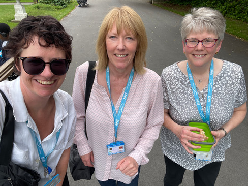 Three people stand on a scenic outdoor path lined with trees and greenery, captured during daylight. The walkway appears to be paved and surrounded by grass, with bare tree branches suggesting a winter or early spring season. In the background, there are more trees and open space, creating a calm, natural setting.