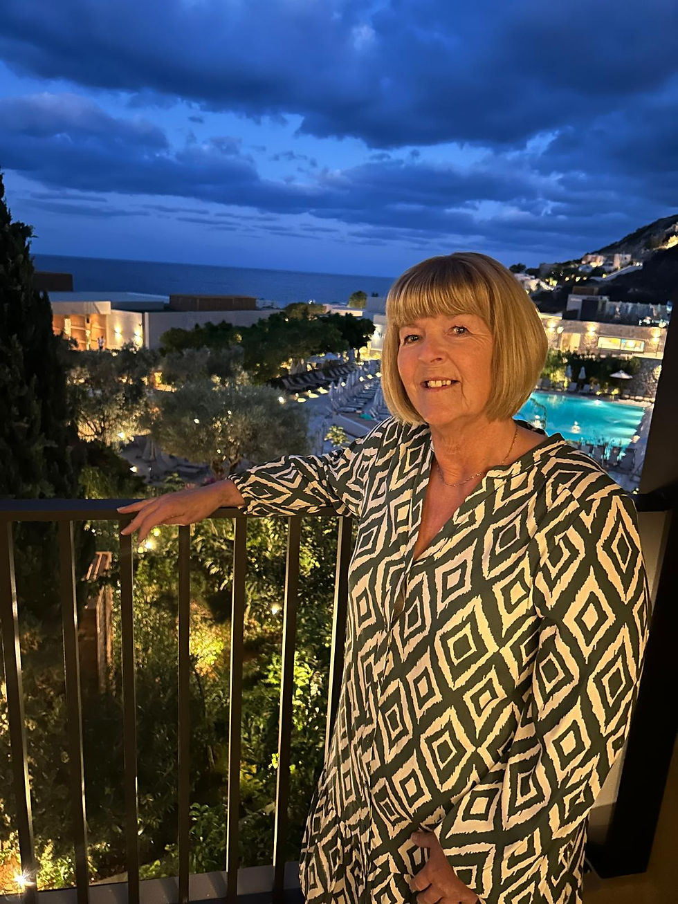 Person standing on a balcony overlooking a coastal resort at dusk. Illuminated gardens and a swimming pool can be seen below, with the sea and dramatic dark clouds stretching across the evening sky. The person is wearing a patterned black-and-white top and resting one arm on the balcony railing.