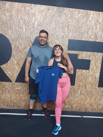 Smiling man and woman in a gym pose with a blue shirt in front of a plywood wall with bold black lettering.
