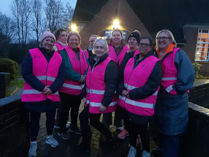 A group of people wearing bright pink high-visibility vests stand together on a paved path outdoors at dusk. The group is gathered in front of a brick building with lit windows and a sign that reads “Holy Nativity Church.” Trees and a low brick wall surround the area, and several people are dressed in coats, hats, and trainers.