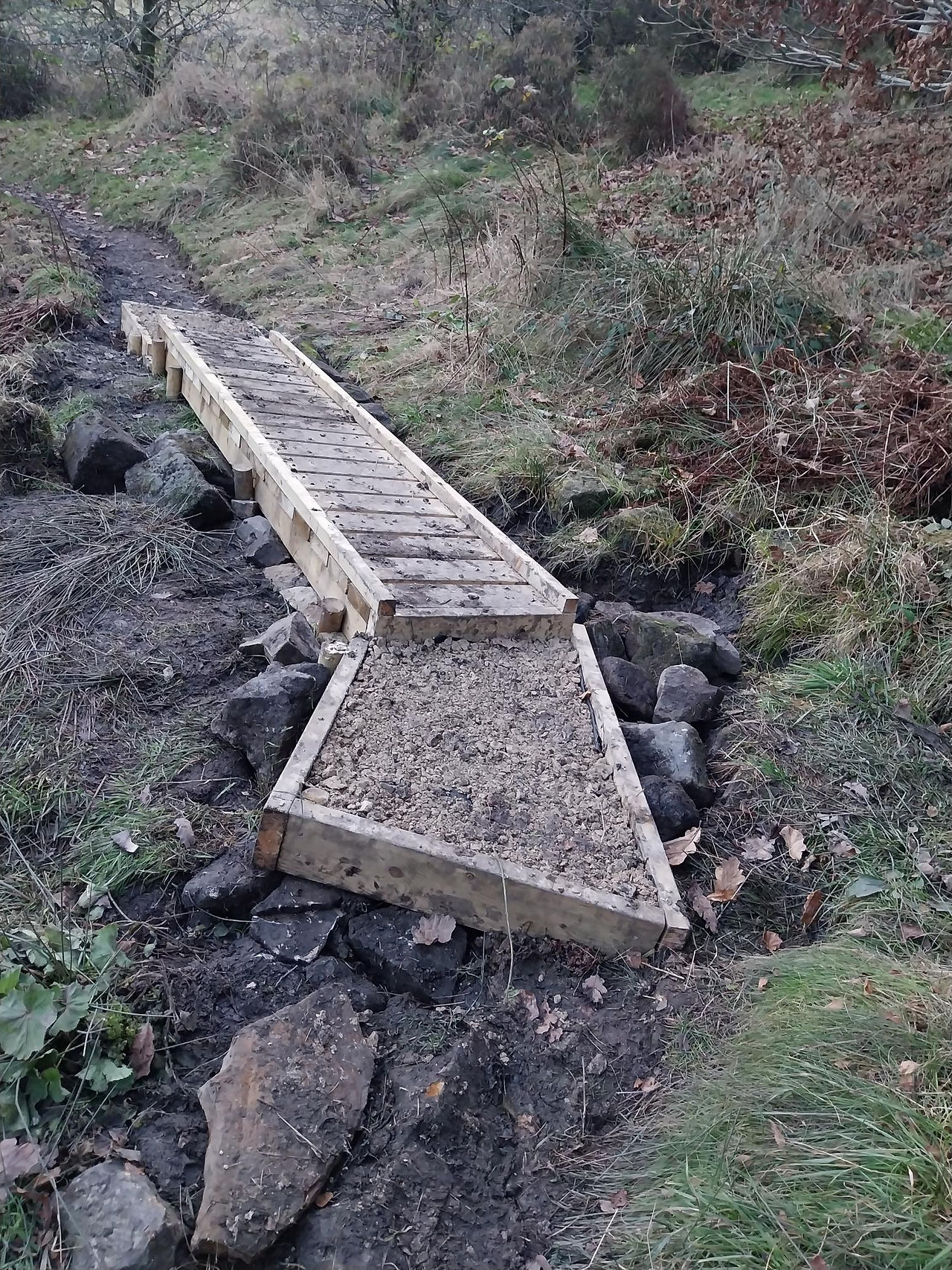 A newly built wooden boardwalk winding through a muddy, grassy hillside, providing a clear walking path. Bare trees with autumn leaves surround the area, and the path curves gently uphill into the distance.