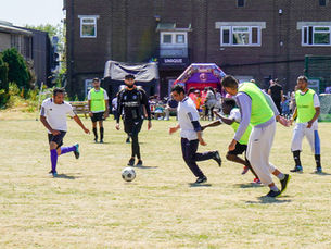 Men playing soccer on a sunny grassy field, with neon bibs and a ball near center; tents and a brick building labeled UNIQUE behind.