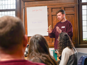 Man in a maroon sweater speaks to an audience, gesturing with a notepad. A flip chart reads "Youth Sport Trust, Rounders England."