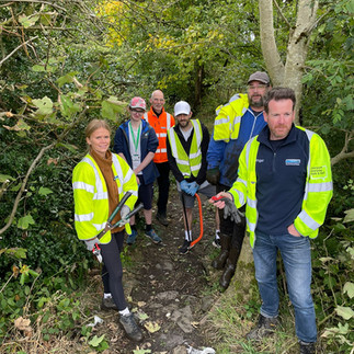 A group of seven people wearing high-visibility jackets and safety gear are standing on a narrow, rocky path surrounded by dense greenery. Some individuals are holding tools such as saws and pruners, indicating they might be engaged in a conservation or maintenance activity. The background shows trees and bushes, suggesting the location is a wooded area.