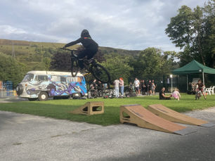 A cyclist in black clothing and helmet performs a jump on a BMX bike over wooden ramps at an outdoor event. In the background, people watch near a green gazebo and a van decorated with colorful graffiti, with hills and trees under a cloudy sky.