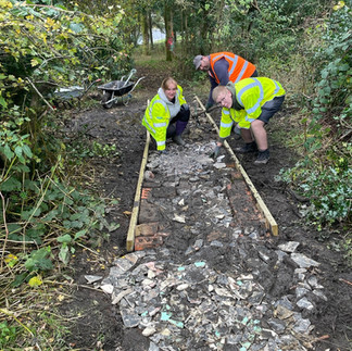 Three individuals wearing high-visibility jackets are constructing a pathway in a wooded area. They are placing broken pieces of tiles or stones within a wooden frame that outlines the path. A wheelbarrow is visible in the background, and the surrounding area is dense with greenery and trees.