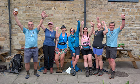A group of eight walkers standing in front of an old stone wall outside, raising their arms in celebration. Several people are wearing medals around their necks and hiking gear, including boots and backpacks. One person on the left is holding up a drink. Wooden picnic benches and bags are visible behind and beside the group, suggesting the end of a group walk or achievement celebration.
