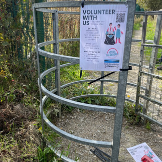 A metal structure with curved bars is placed on a gravel path surrounded by greenery. Attached to the structure is a poster that reads 'VOLUNTEER WITH US' and includes details about a community clean-up event, contact information, and a QR code. Another similar poster lies on the ground nearby. In the background, there is a fenced area with trees and a partly cloudy sky