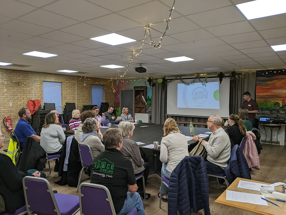 A group of people sit around several tables arranged in a large community hall, watching a presentation projected onto a screen at the front of the room. The screen displays the title ‘Active Rastrick’. String lights are draped across the ceiling, and the room has brick walls, purple chairs, and murals painted on the back walls. Papers, notebooks, and cups are on the tables as attendees listen to the presenter standing near the screen.