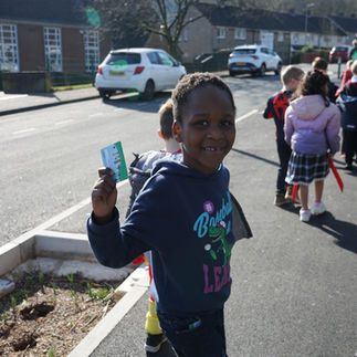 A group of children walking on a sidewalk next to a road. One child in the foreground, wearing a dark blue hoodie with colorful designs, is holding up a small card. The child's face is obscured for privacy. Other children are visible in the background, some wearing backpacks and coats. There are houses and parked cars along the road. The weather appears sunny with clear skies.