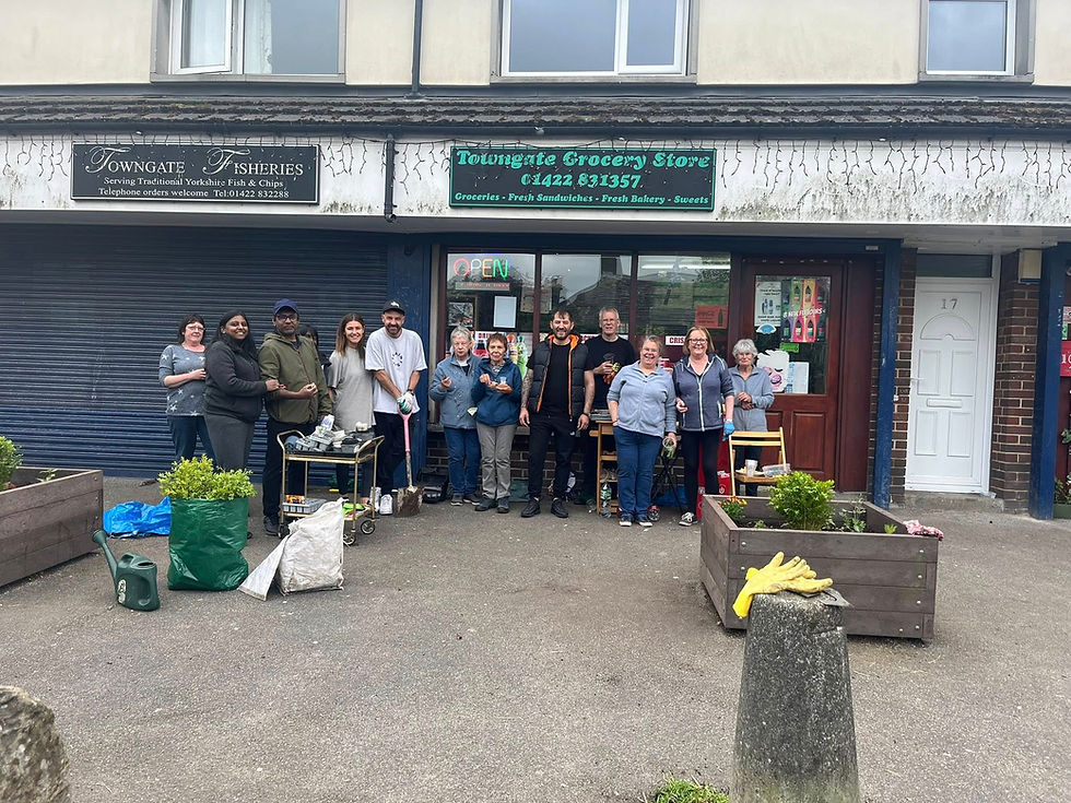 A group of people standing in front of a storefront. The storefront has two signs above it, one reading "Towngate Fisheries" and the other "Towngate Grocery Store." The group appears to be posing for a photo, with some individuals holding gardening tools and supplies. There are large planters with greenery on either side of the entrance. The store's address is 17, visible on the door to the right.