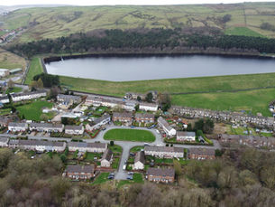 An aerial view of a residential neighborhood with rows of houses, winding roads, and green lawns. In the background, there is a large rectangular reservoir surrounded by grassy embankments and dense forested areas. Beyond the reservoir, rolling hills with patches of farmland are visible. The image captures a mix of urban and natural landscapes.