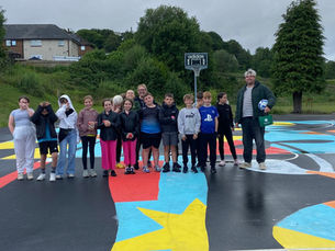 A group of people, including children and an adult, are standing on a colorful outdoor basketball court decorated with bright patterns and shapes. A basketball hoop with an 'adidas' logo is visible in the background. The setting is a residential area with houses and greenery, under an overcast sky.
