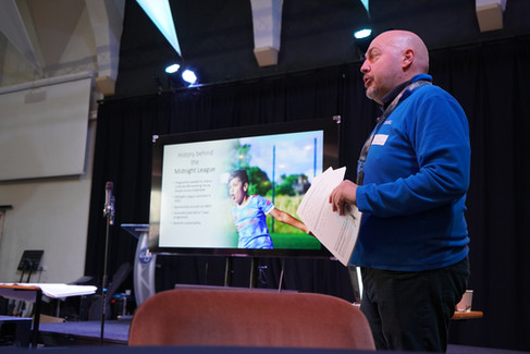 Man in blue sweater presents by a screen showing "History behind the Midnight League" in a conference room with a dark curtain backdrop.
