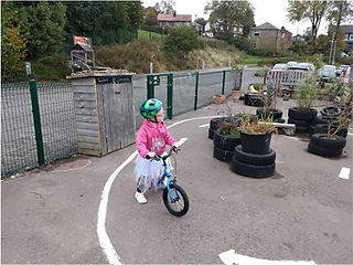 Child in a pink hoodie and tutu rides a blue bike in a playground with tire planters and a shed. Background shows trees and houses.