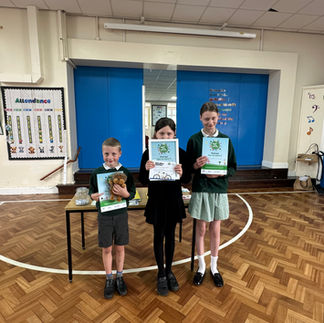 Three children stand in a school gymnasium holding certificates and a stuffed animal. The child on the left holds a certificate and a teddy bear, the child in the middle holds up a certificate with both hands, and the child on the right holds a certificate. Behind them is a table with additional items, blue doors, and an attendance chart on the wall to the left. The floor is wooden parquet with white lines marking sports boundaries.