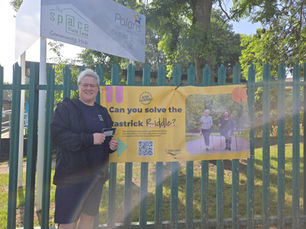 A person stands smiling next to a green metal fence, holding a gift voucher. Behind them is a yellow banner that reads "Can you solve the Rastrick Riddle?" with an image of two people walking in a park. The banner also includes a QR code and logos for Active Calderdale and Calderdale Council. Above the fence, signs for "The Space Field Lane Community Hub" and "Polaris Out-of-School Club" are visible. Trees and greenery can be seen in the background on a sunny day.