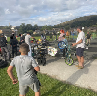 A community event taking place outdoors in a park, with several people gathered around a bike repair or maintenance station. Multiple bicycles are being worked on, one held upside down for repairs. A sign reads "Active Travel Hub." Children and adults are present, some watching and others helping. Behind them are houses, green hills, and a cloudy blue sky. Playground equipment is visible to the right.