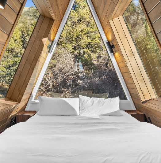 Loft bedroom in an A-frame cabin with large sky windows and a plush white bed overlooking surrounding nature.