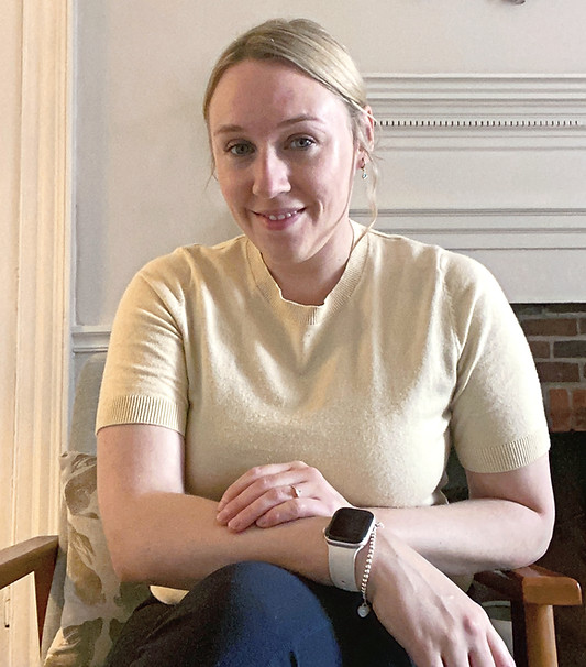 A woman with blonde hair in an up-do is wearing a light yellow jumper with short sleeves, sitting in a chair and smiling