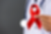 Foto de um médico ao fundo desfocado segurando em primeiro plano o laço vermelho simbolo do combate ao HIV