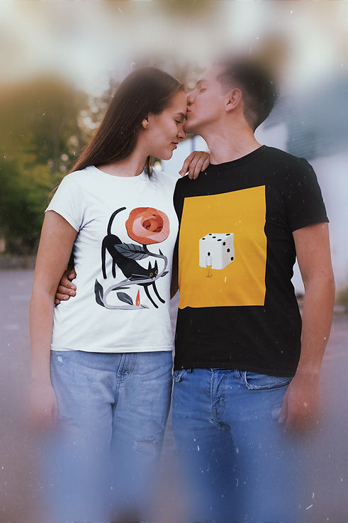 The image features a couple showcasing artistic T-shirts from the AGLGartwear collection. The woman wears a white T-shirt with a design of a black cat, a stylized rose, and a red droplet, highlighting the creativity and storytelling behind the art. The man wears a black T-shirt with a conceptual design of a large white die on a yellow background, accompanied by a small human figure, emphasizing the surreal and thought-provoking nature of the artwork. Both T-shirts are part of a unique collection that transforms fashion into wearable art, celebrating individuality and creativity. The setting suggests a relaxed and intimate outdoor moment.