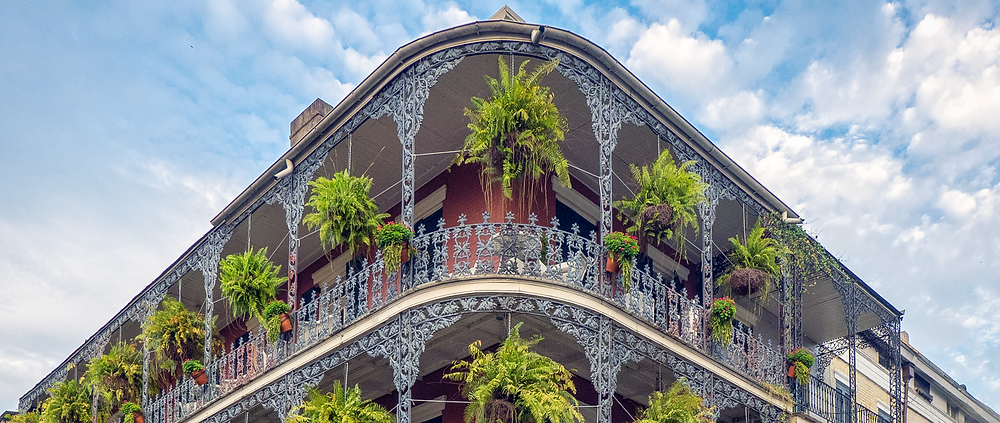 The corner of a building with a wrap-around decorative balcony and ferns in hanging planters.