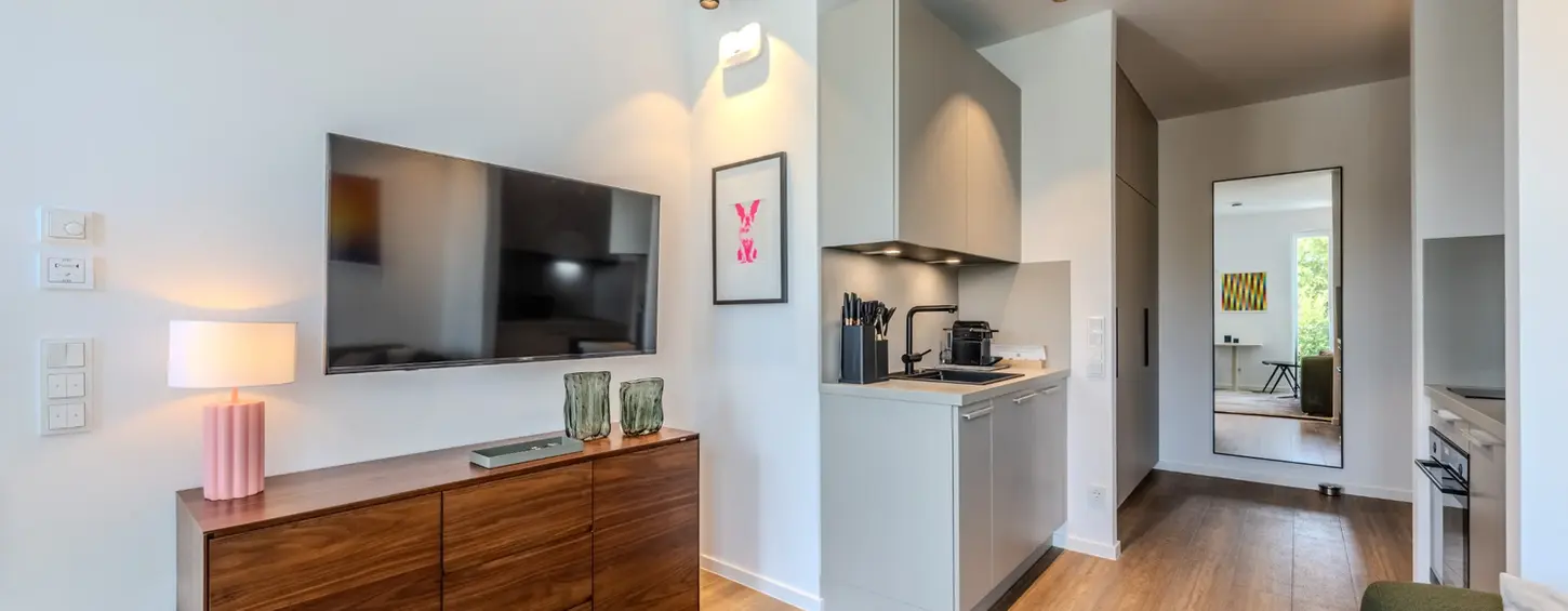Wohnbereich mit TV-Konsole und Blick in die Küche in einem vollständig möblierten Studio M Apartment