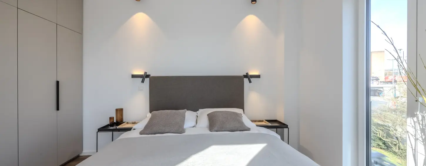 Schlafzimmer mit Wandleseleuchten und großem Fenster in einem vollständig möblierten Apartment XL