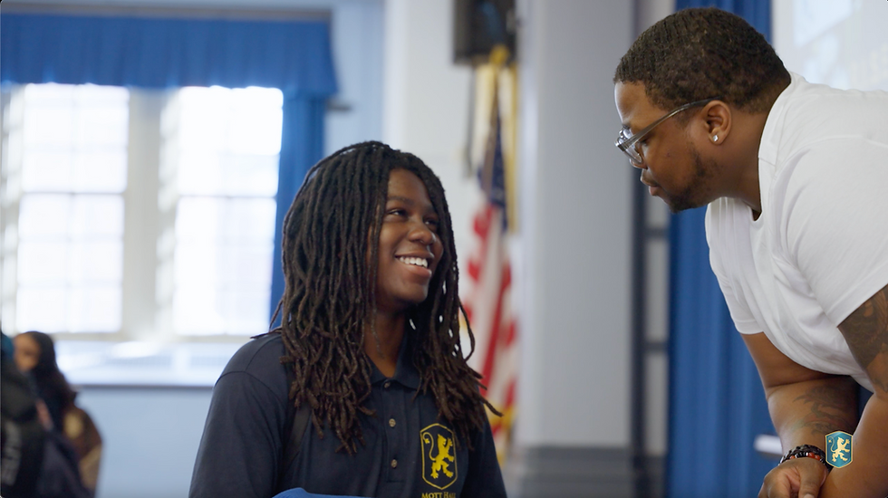A smiling student wearing a Mott Hall school uniform polo shirt shares a positive one-on-one moment with an adult mentor or staff member in a school common area. An American flag is visible in the background, and the Mott Hall school logo appears in the corner of the image.