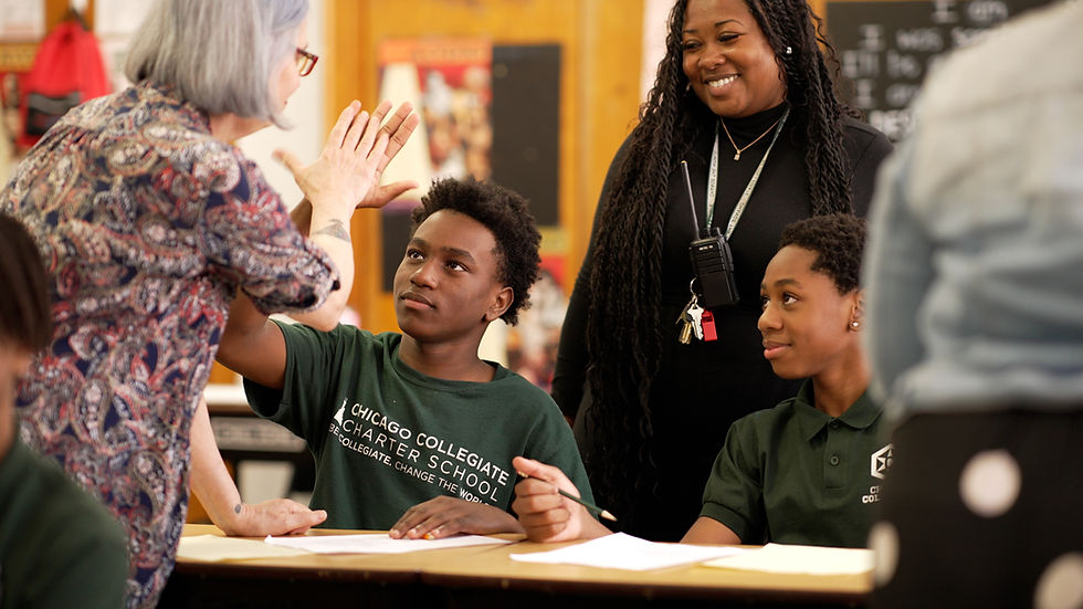 A middle school student wearing a Chicago Collegiate Charter School t-shirt raises his hand to high-five a teacher during a classroom lesson, while a smiling adult staff member looks on and a fellow student sits nearby. The scene captures an engaged and supportive learning environment at Chicago Collegiate Charter School.
