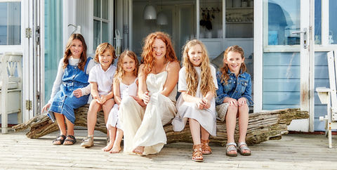 Six children sitting on driftwood, styled in light white and denim clothing, as part of a summer campaign image