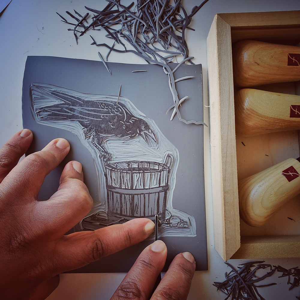 Marta's black hands holding a carving instrument to the surface of a lino plate with an image of a crow, wash bucket and bubbles. The image is half carved. There are lino plate shavings to the side of the plate. The right side of the image shows handles of additional carving tools in a wooden box