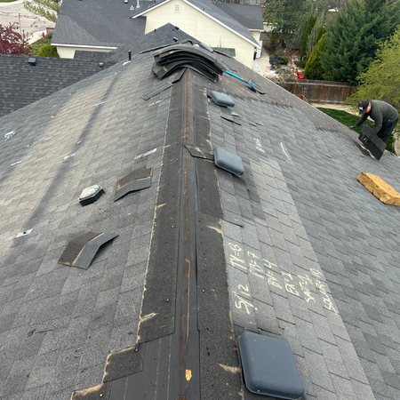A high-angle aerial photograph of a roof under construction. Workers are actively removing old shingles. Sections of exposed brown OSB decking are visible alongside existing grey shingles that are yet to be removed. Debris and tools are scattered on the roof.