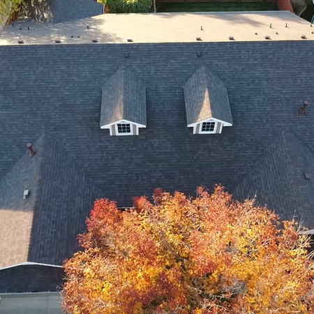 A high-angle aerial view of the completed project. The home features new, dark charcoal architectural shingles. Two dormer windows with crisp white trim stand out against the clean, uniform shingle lines.