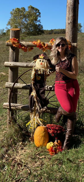 Woman poses with a scarecrow, pumpkins, and a rustic wheel for fall.