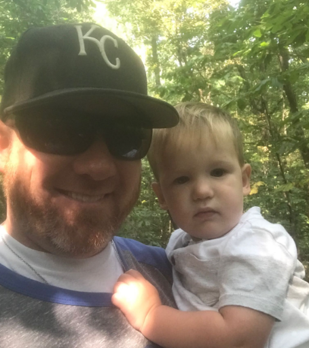 Man wearing KC hat and sunglasses holding a young child in nature.
