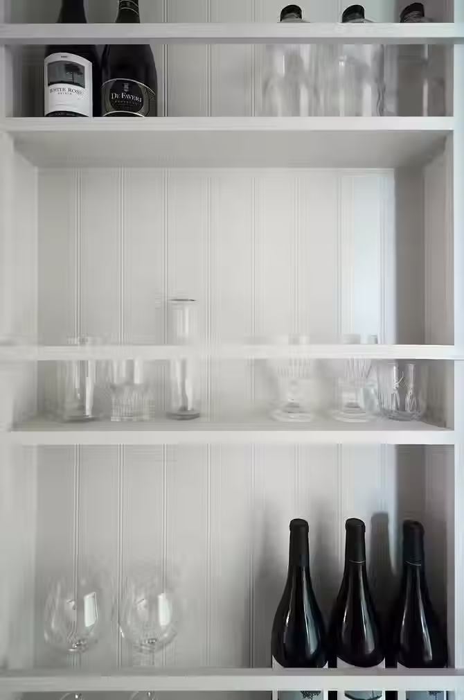 A close up of the built in cabinetry inside of a walkway for glassware and wine. There are wine glasses and wine bottles displayed inside the white painted wooden paneled cabinetry.