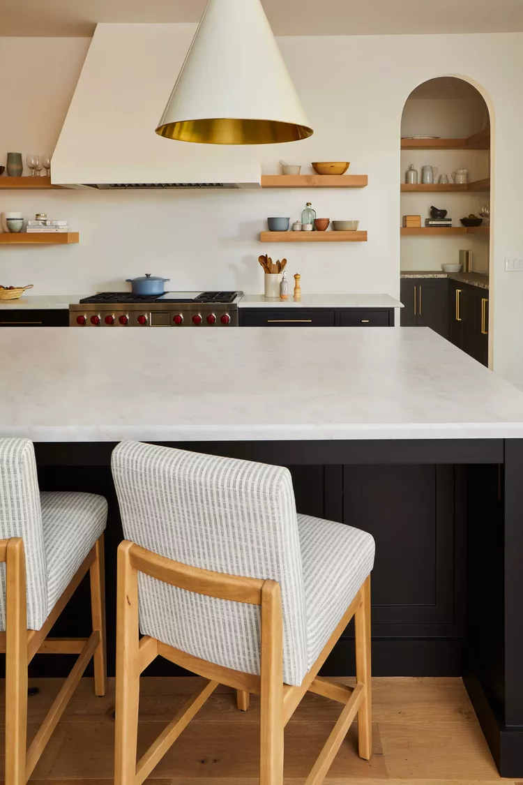 Modern kitchen with a white island, formica countertops, striped chairs, open wooden shelves, pendant light, and stove with blue pot. Calm and organized decor.