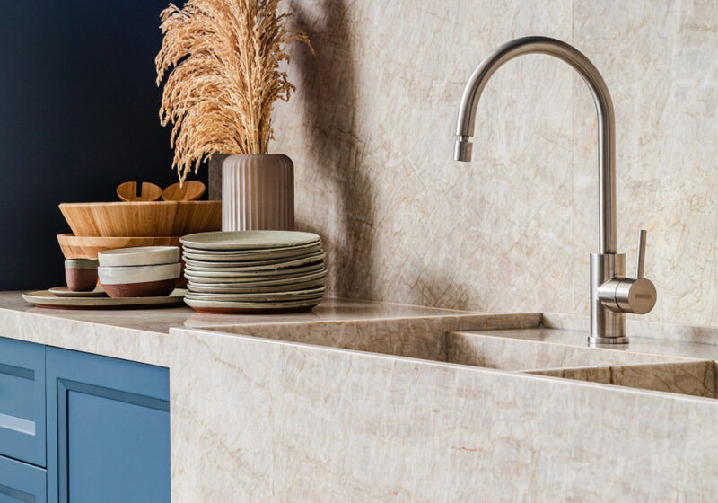 A unique looking quartzite countertop and sink that is sand colored and on the backsplash as well. There is dish ware stacked next to the sink along with a vase. There are blue cabinet drawers below.
