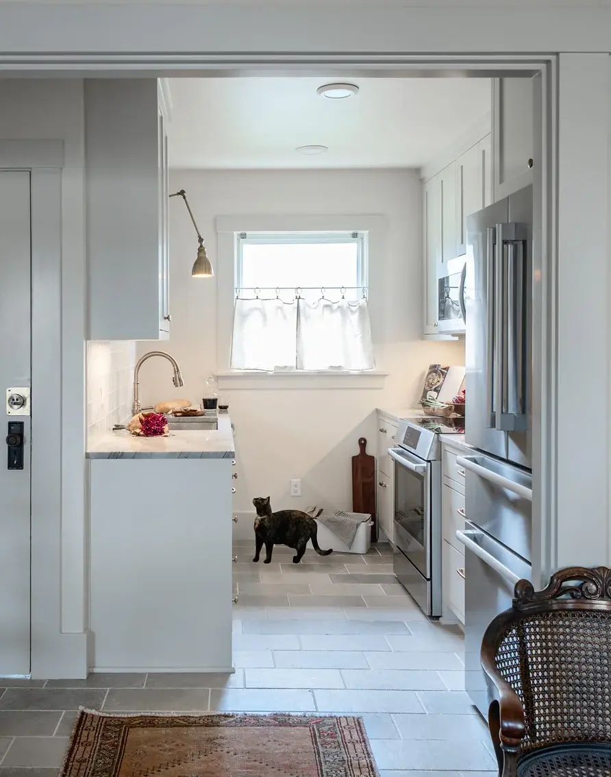 A kitchen nook with warm task lighting and a small window. The room is all neutrals with tile flooring and a cat looking up at the counter.