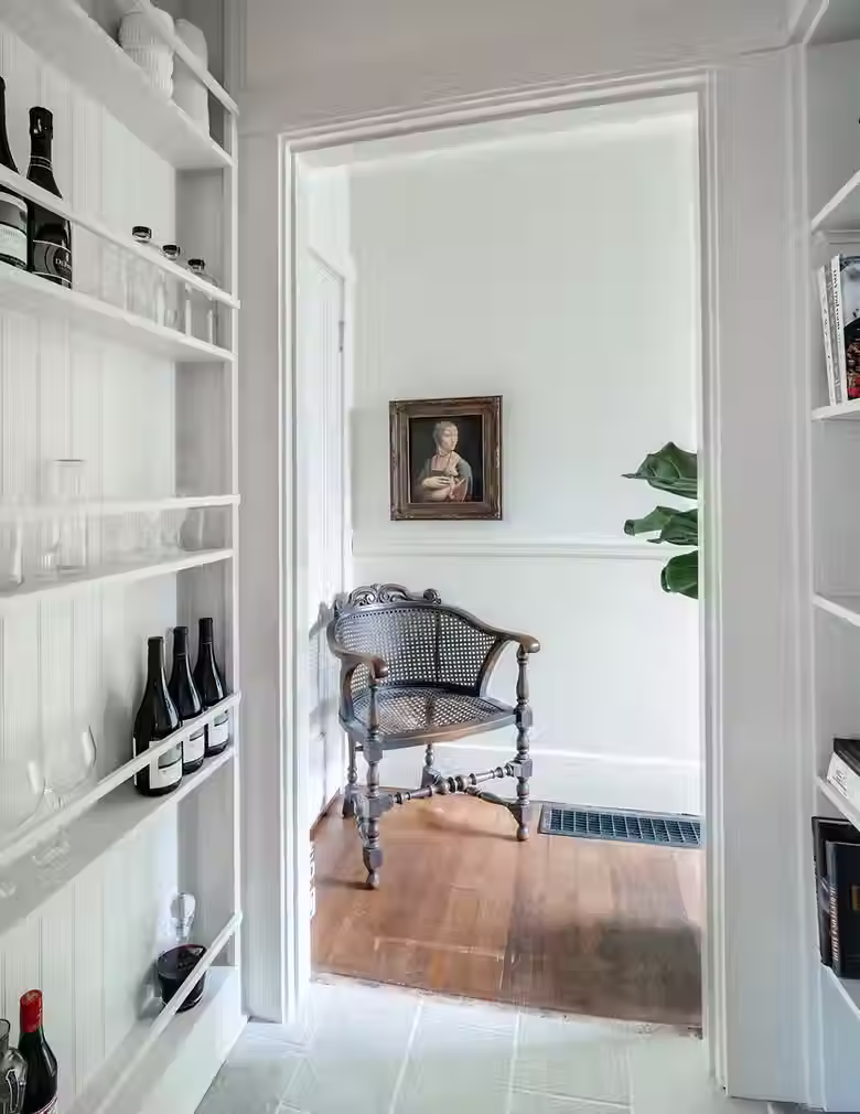 A walkway with built in cabinetry for glassware. The walk way is paneled with white painted wood and it leads to a room with an antique chair and painting hanging above it.