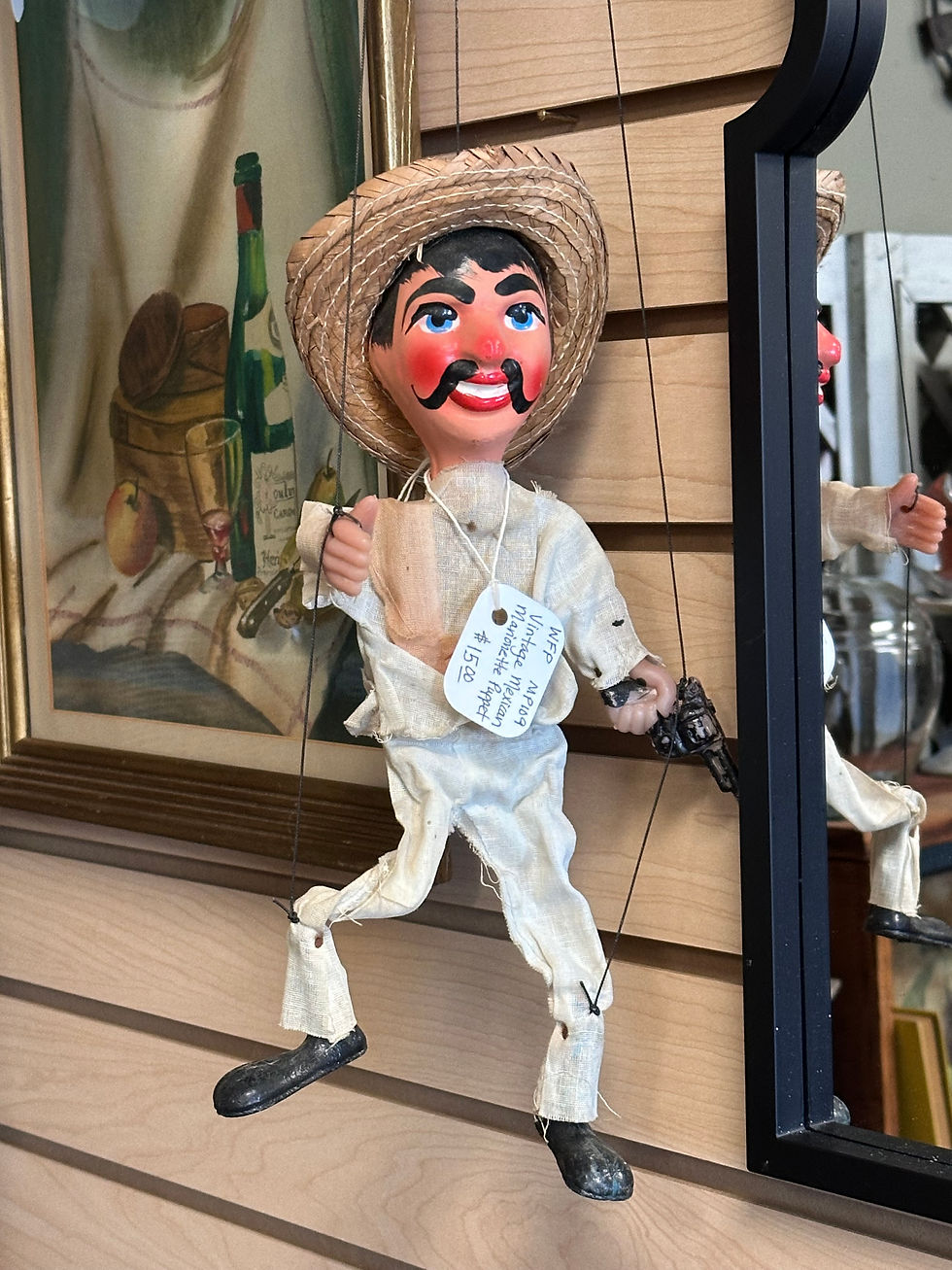 a marionette of a Spanish or Mexican man; his face is painted flushed, with a black mustache and a wide smile. He wears a straw hat; one of his hands is open while the other holds a gun. His body is limp and flat, except for black shoes that are well worn. He's held together with four black strings.