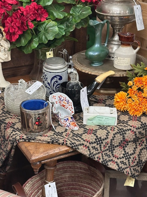 A table draped with a geometric tablecloth, filled with various vintage items, such as a stamp roller, fake marigolds, a beer stein, and glassware plates.