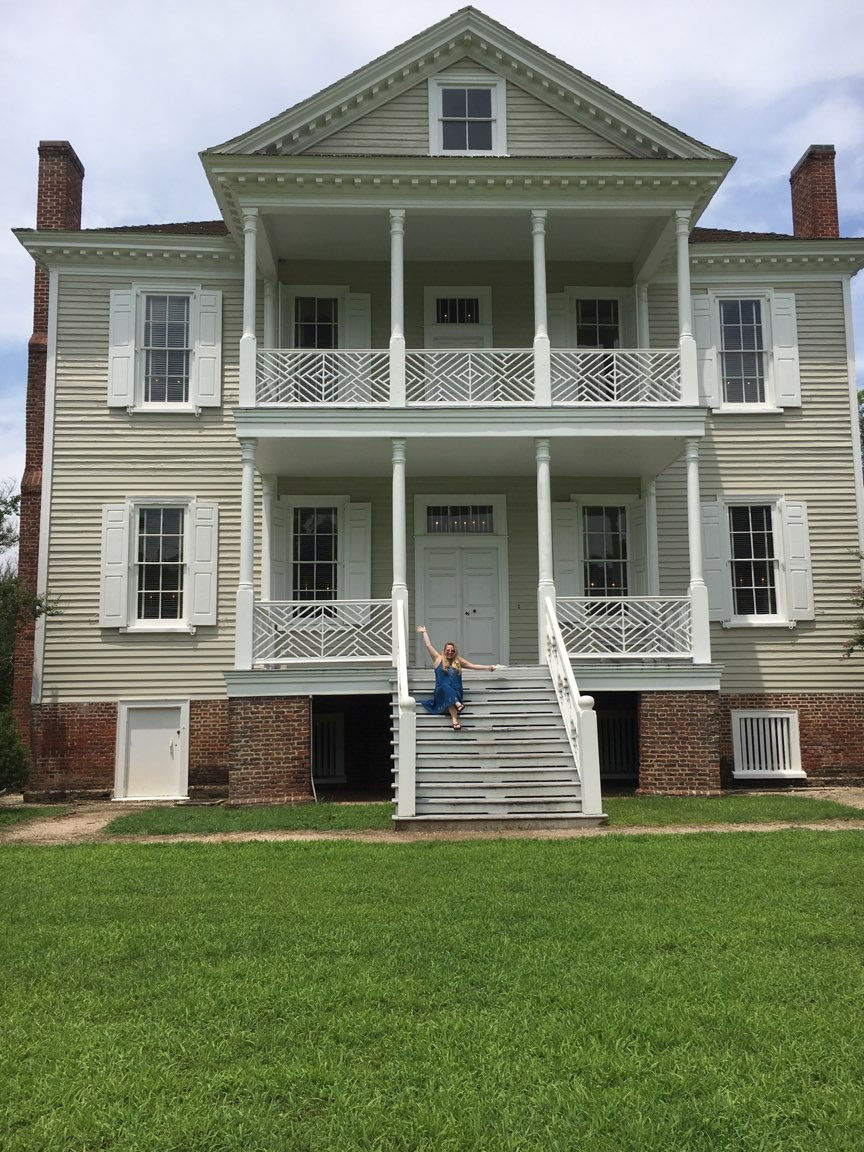 The front of an 18th Century home called The Hope House. A blonde woman sits on the front steps in a deep blue dress, with her arms out to the sides.