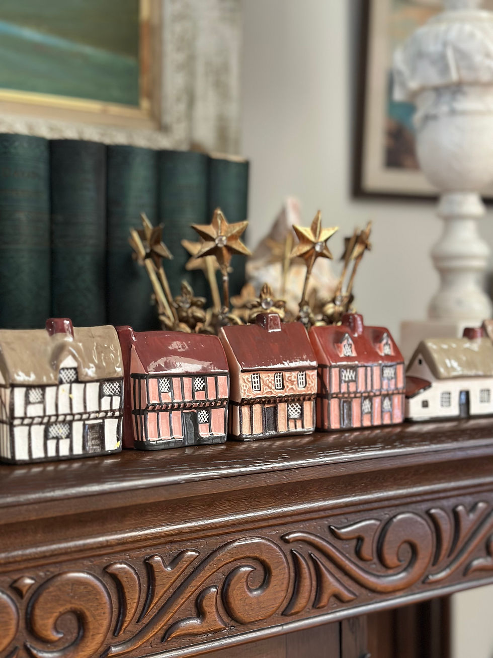 A close-up photo of red-and-brown Mudlen End Cottages on the hand carved wooden shelf with the antique golden crown behind it; there are green books and a marble lamp right beside them.