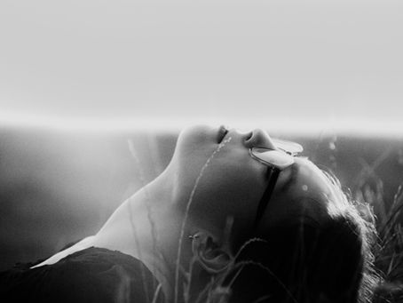 Person lying in tall grass with eyes closed and glasses catching the light, shown in side profile.