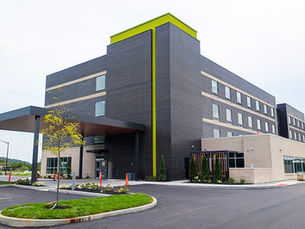 Modern dark-brick hotel with lime-green trim and covered entrance, empty driveway, under an overcast sky.