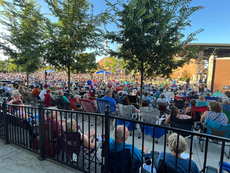 Large crowd seated in folding chairs outdoors under trees, watching a stage at a sunny community concert.
