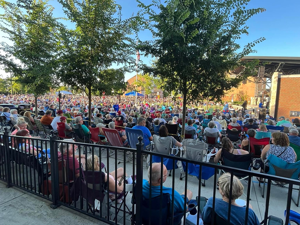 Large crowd seated in folding chairs outdoors under trees, watching a stage at a sunny community concert.